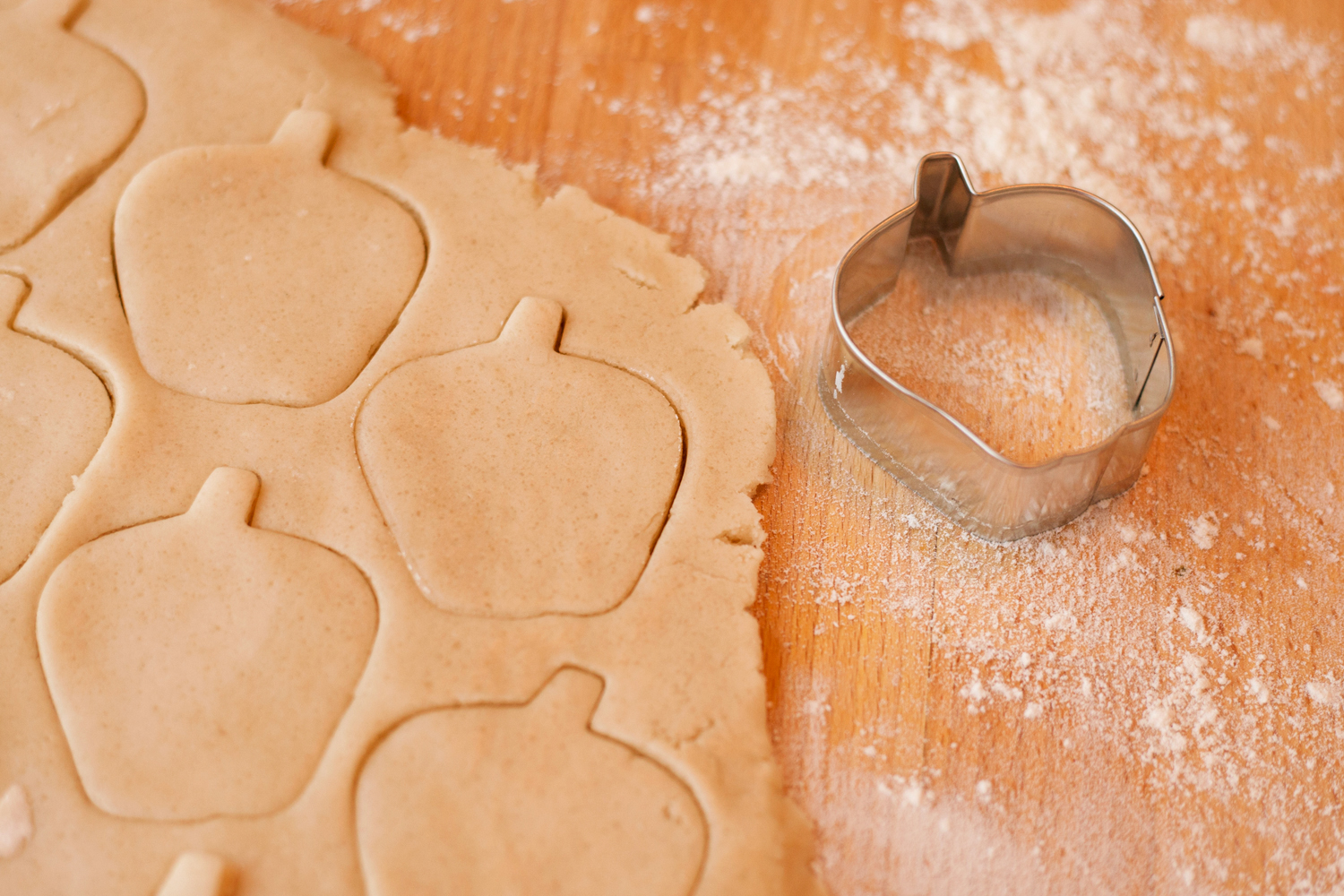 An apple-shaped metal cookie cutter beside rolled dough with several apple shapes cut into it